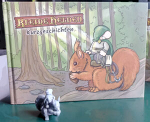 Buch „Kleine Helden – Kurzgeschichten“, Hardcover. Auf dem gezeichneten Cover ist eine Gestalt mit grünem Umhang und Knollennase zu sehen, die auf einem Eichhorn mit Nuss in den Pfoten durch den Wald reitet. Davor steht eine graue Kunststofffigur des Eichhornreiters auf einer grünen Tischplatte.
